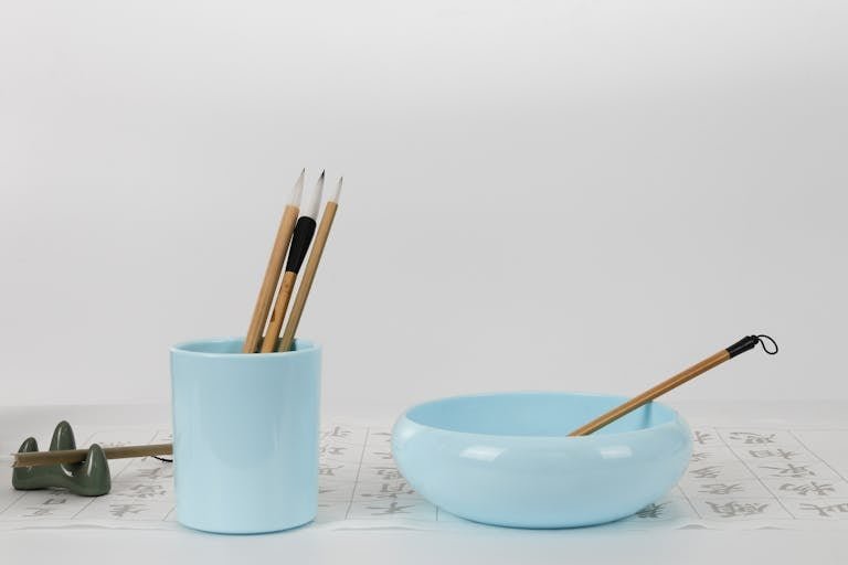 Sophisticated setup of calligraphy brushes in a light blue ceramic holder and bowl on white background.